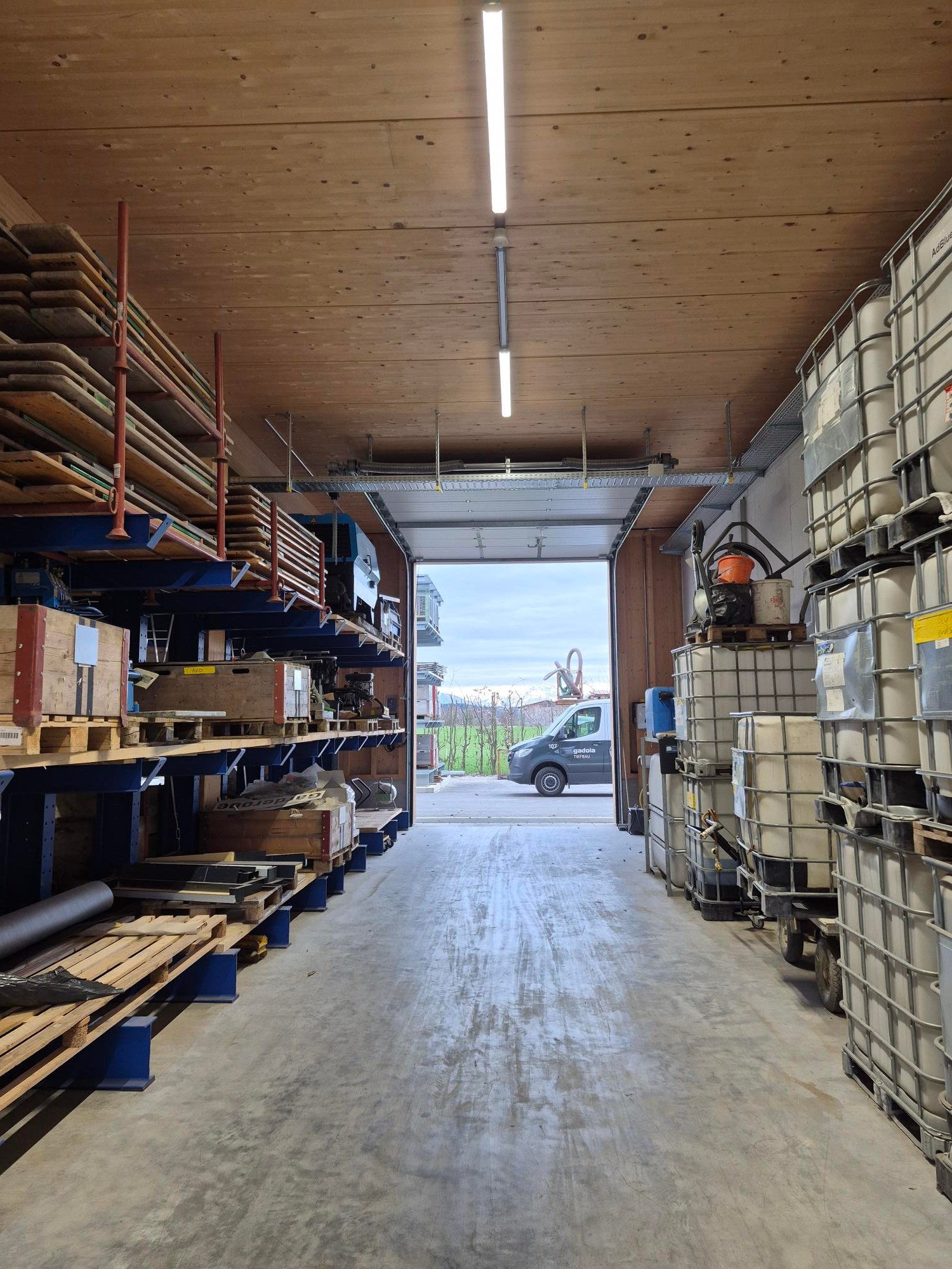 Warehouse with wooden shelves, metal racks, and storage boxes. Open door leading to a road with a van parked outside.