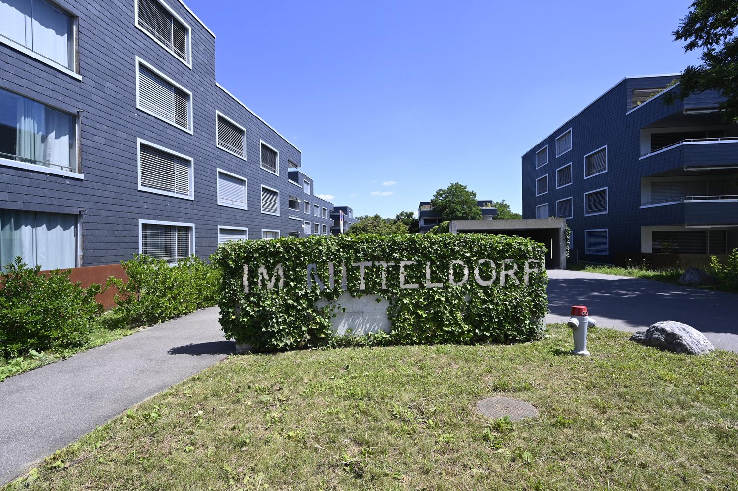 Modern apartment building with blue exterior, multiple windows, and a sign reading 'IM MITTELDORF'. The building is surrounded by greenery and has a driveway.
