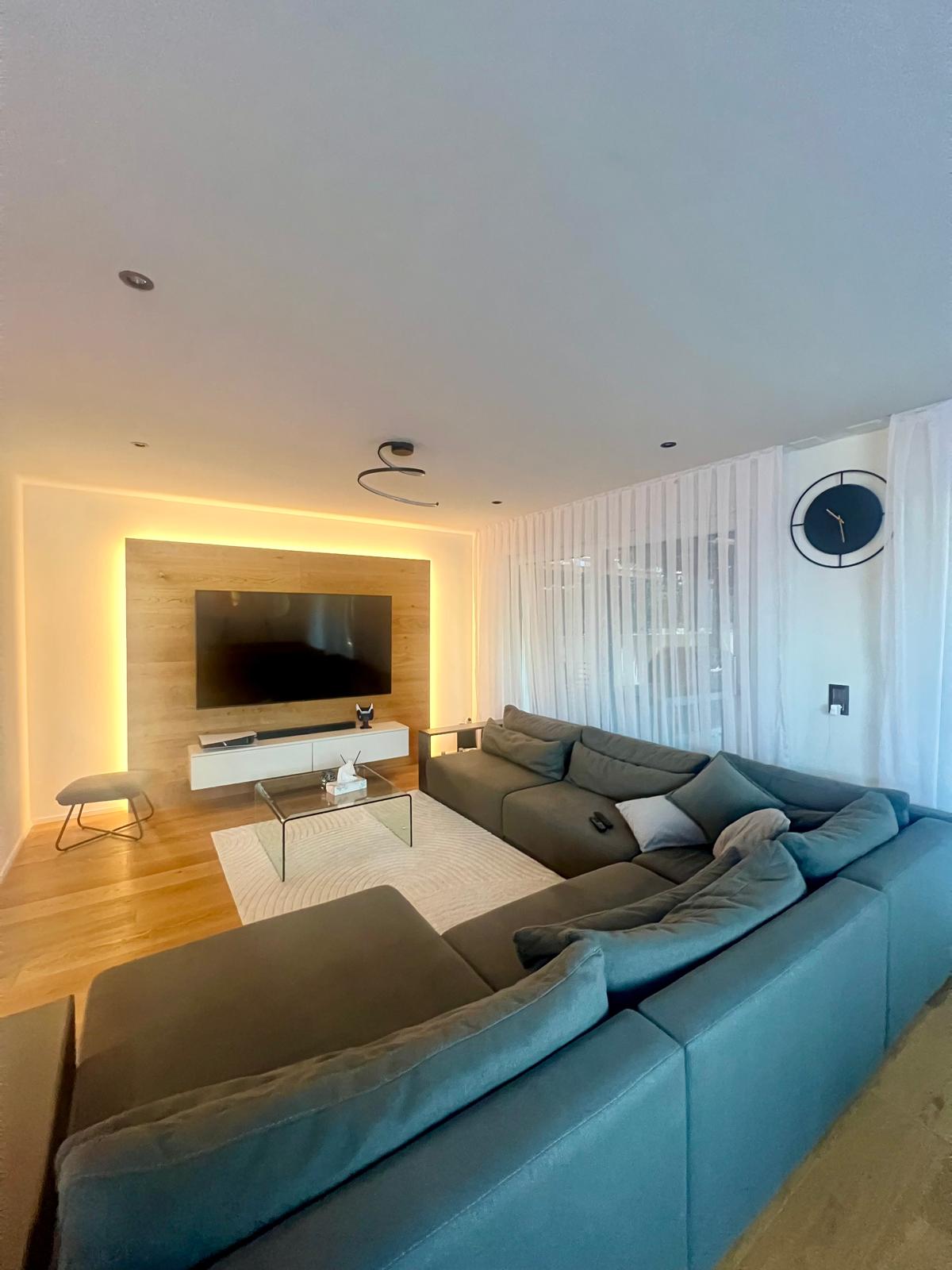 large L-shaped couch, flatscreen TV, glass coffee table, wooden floors, accent lighting, stool, wooden accent wall