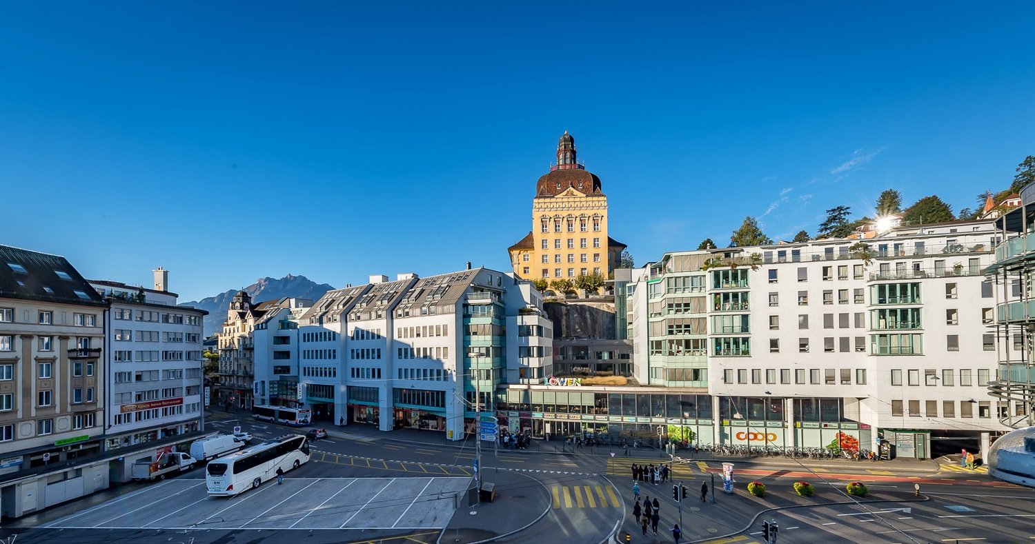 Multiple multi-story buildings with various architectural styles, some with signage, in a downtown area. A clock tower is visible, and in the foreground, a busy intersection with pedestrians and vehicles. The sky is clear and blue.