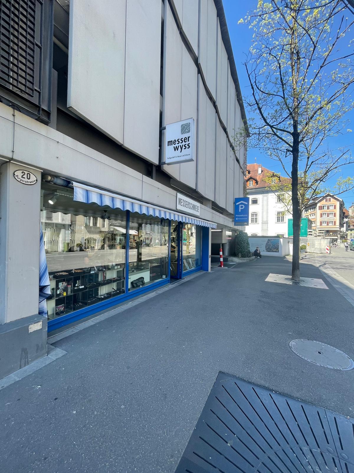 Commercial building with a shopfront, blue awning, and a sign reading 'Messer Wyss'. The building has a modern design with a white exterior and blue accents. The shopfront features large glass windows displaying items inside. There is a parking sign and a