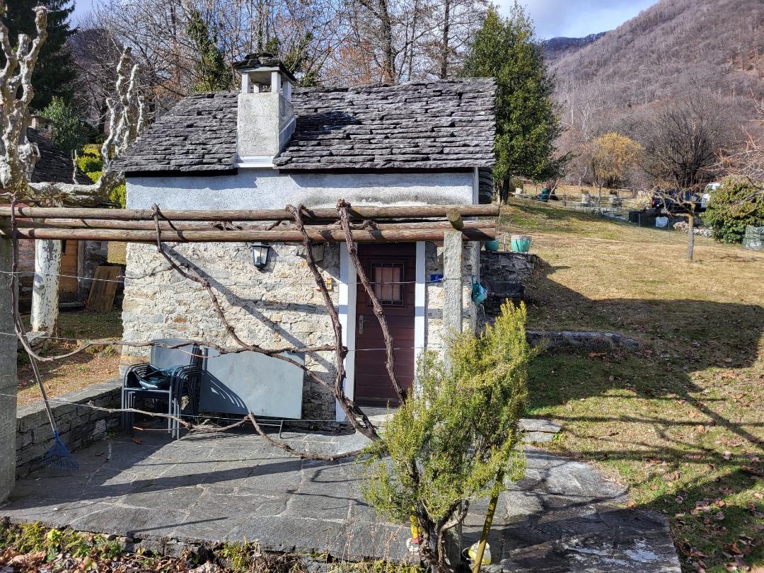 Stone house with chimney, brown door, stone pathway, wooden fence, grassy area, tree, plants, distant mountain