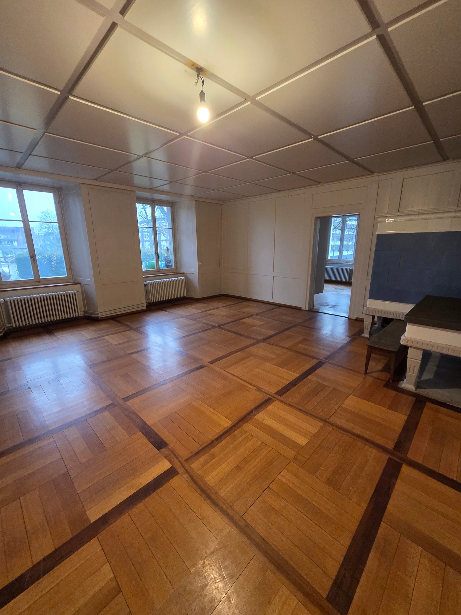 wide room, parquet flooring, white walls, two windows, two radiators, a bench