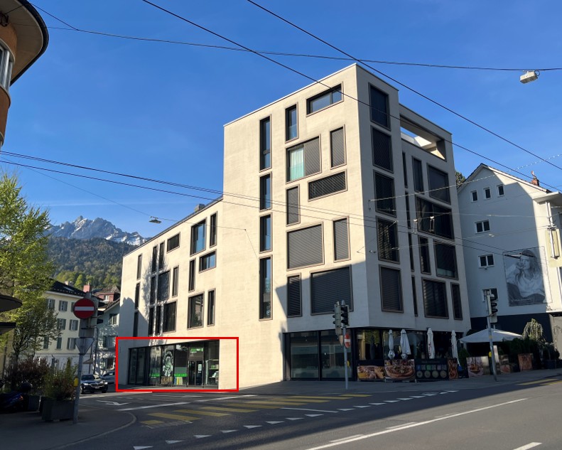 Modern multi-story building with glass facade, located on a street with traffic signs and a pedestrian crossing, surrounded by other buildings and greenery.