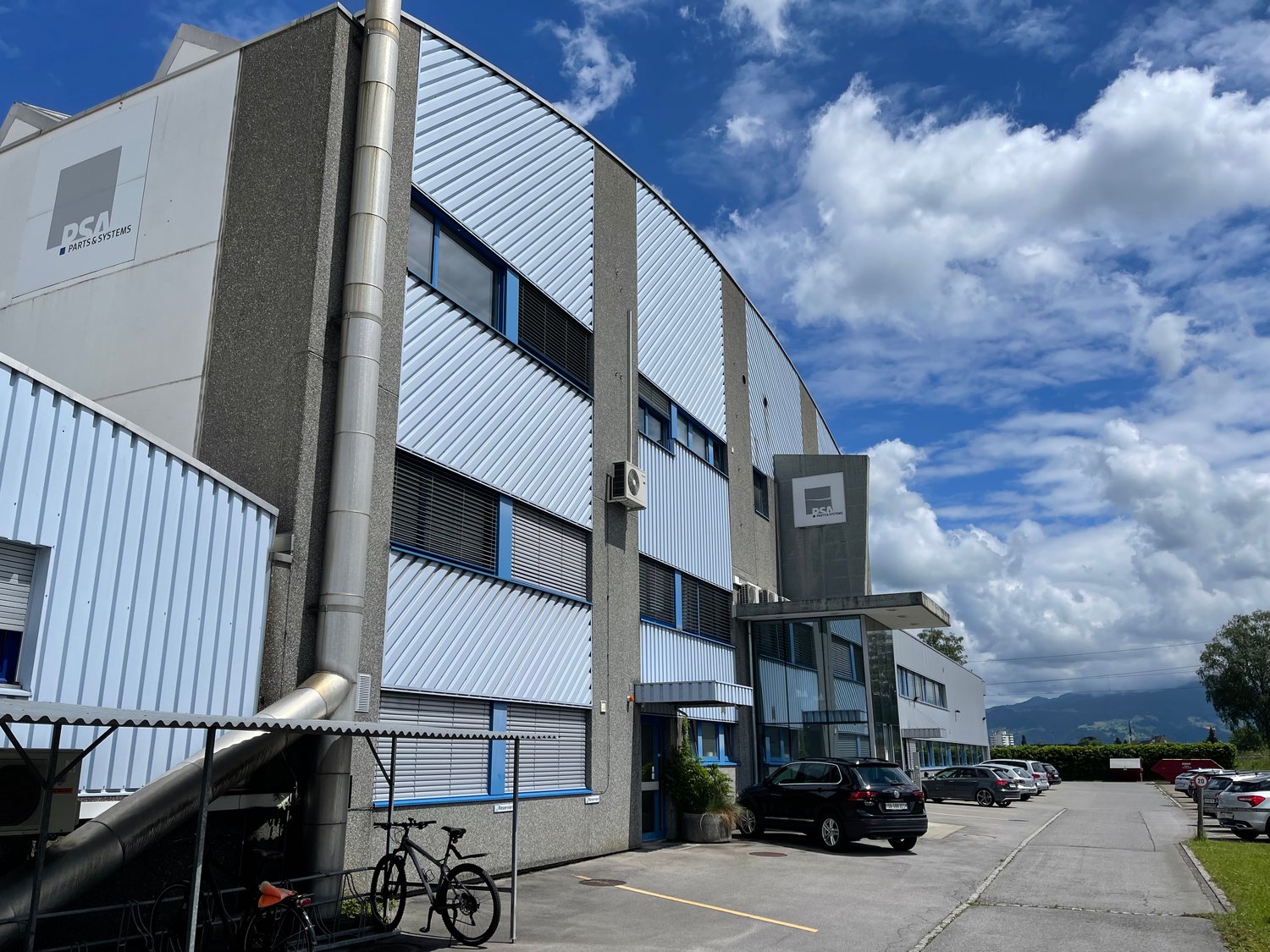 A large industrial building with a distinctive curved roof and metal siding. The building has several windows and a parking lot in front with several cars parked. There is also a bicycle visible in the foreground.
