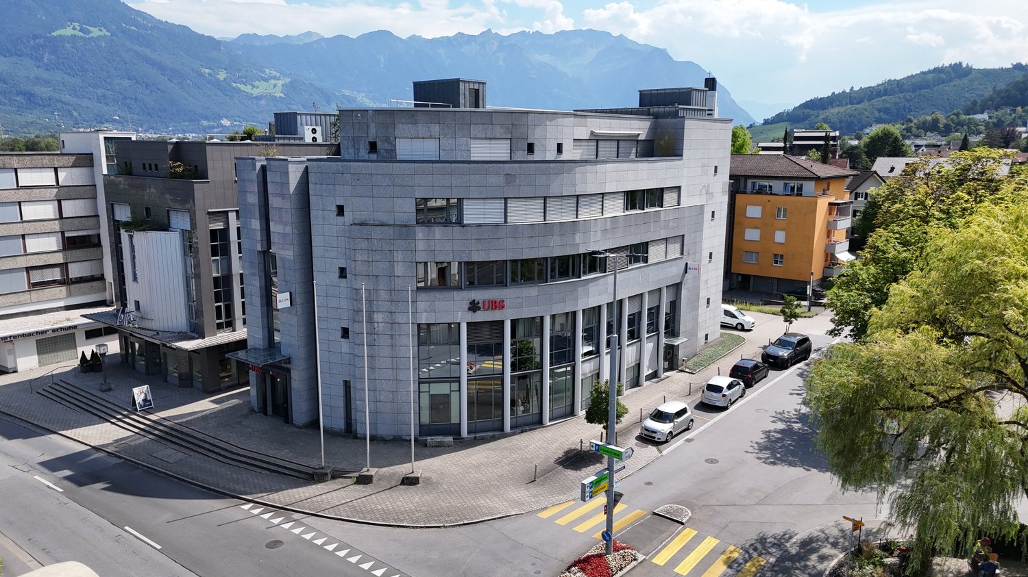 Large modern office building with concrete exterior, multiple floors, and parking spaces in front of the building. The building has the UBS logo displayed on the facade, indicating it is likely an office for the UBS financial services company. The surroun