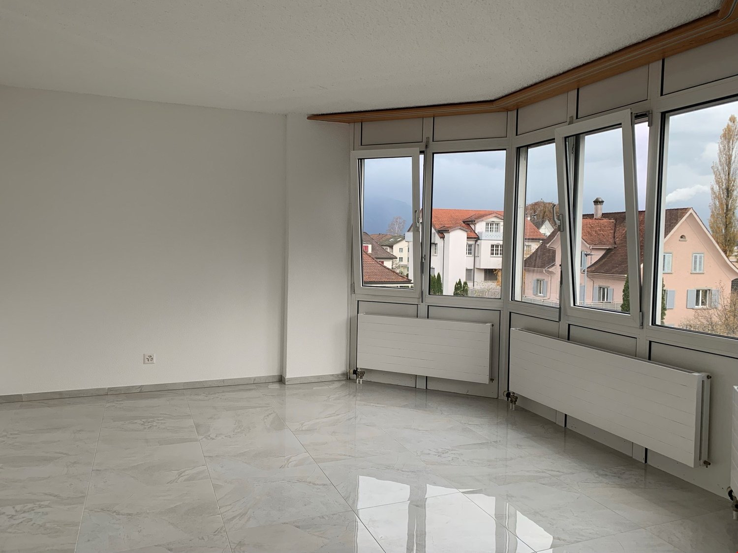 Spacious living room with large windows offering a view of the surrounding buildings, hardwood floors, and a modern, minimalist design.