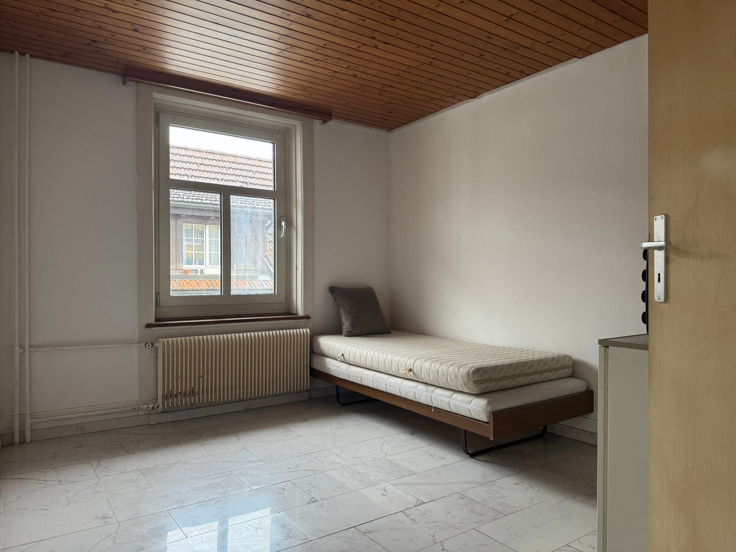 Empty room with a single bed, a window, a radiator, and a wooden ceiling.