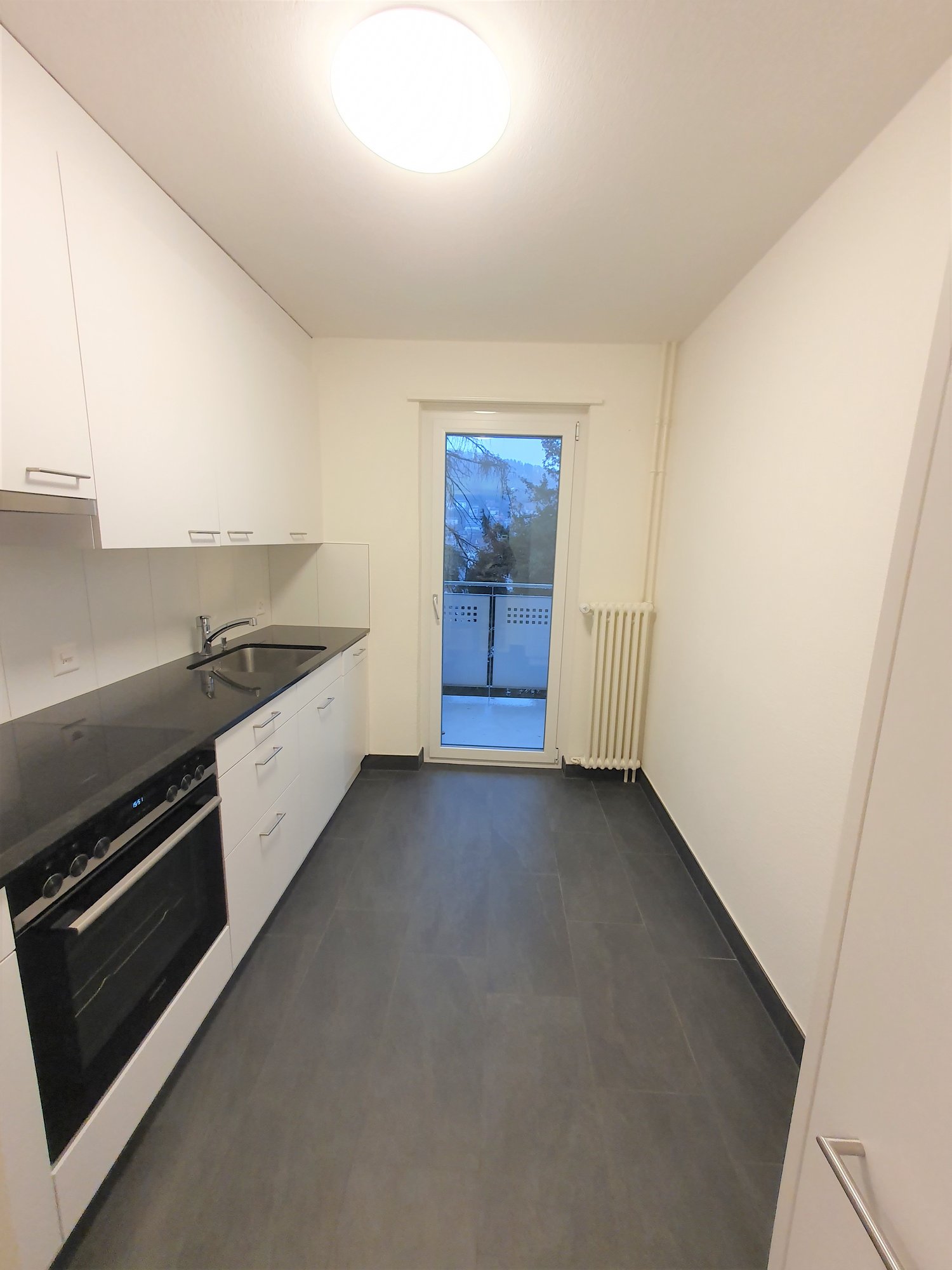 modern kitchen with white cabinets, black countertops, stainless steel sink, oven, and a sliding door leading to a balcony