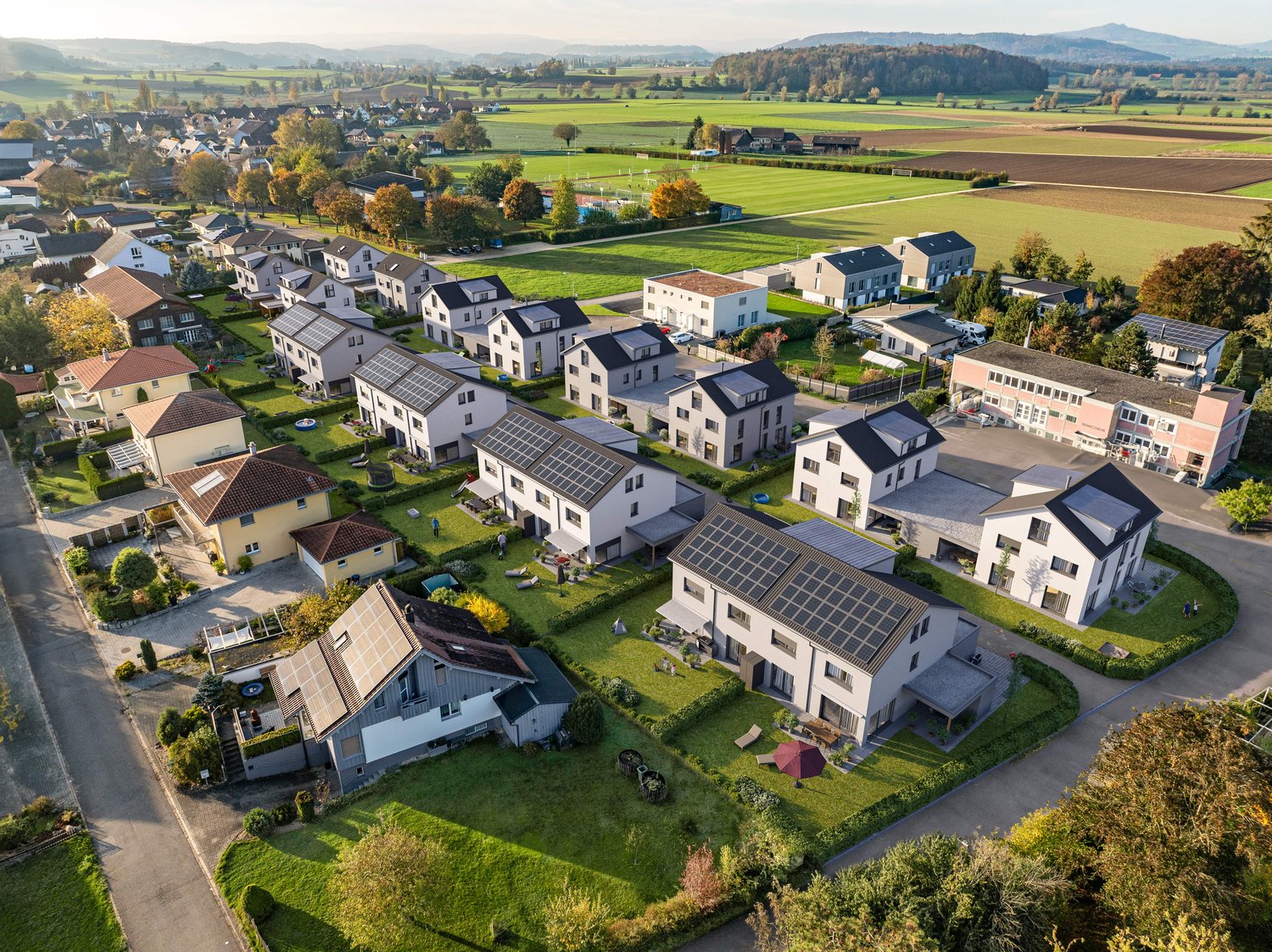 Aerial view of a residential neighborhood with multiple two-story houses, some with solar panels on the roofs. The houses have small yards and gardens, and there are paved parking areas between the buildings. The neighborhood is surrounded by open fields