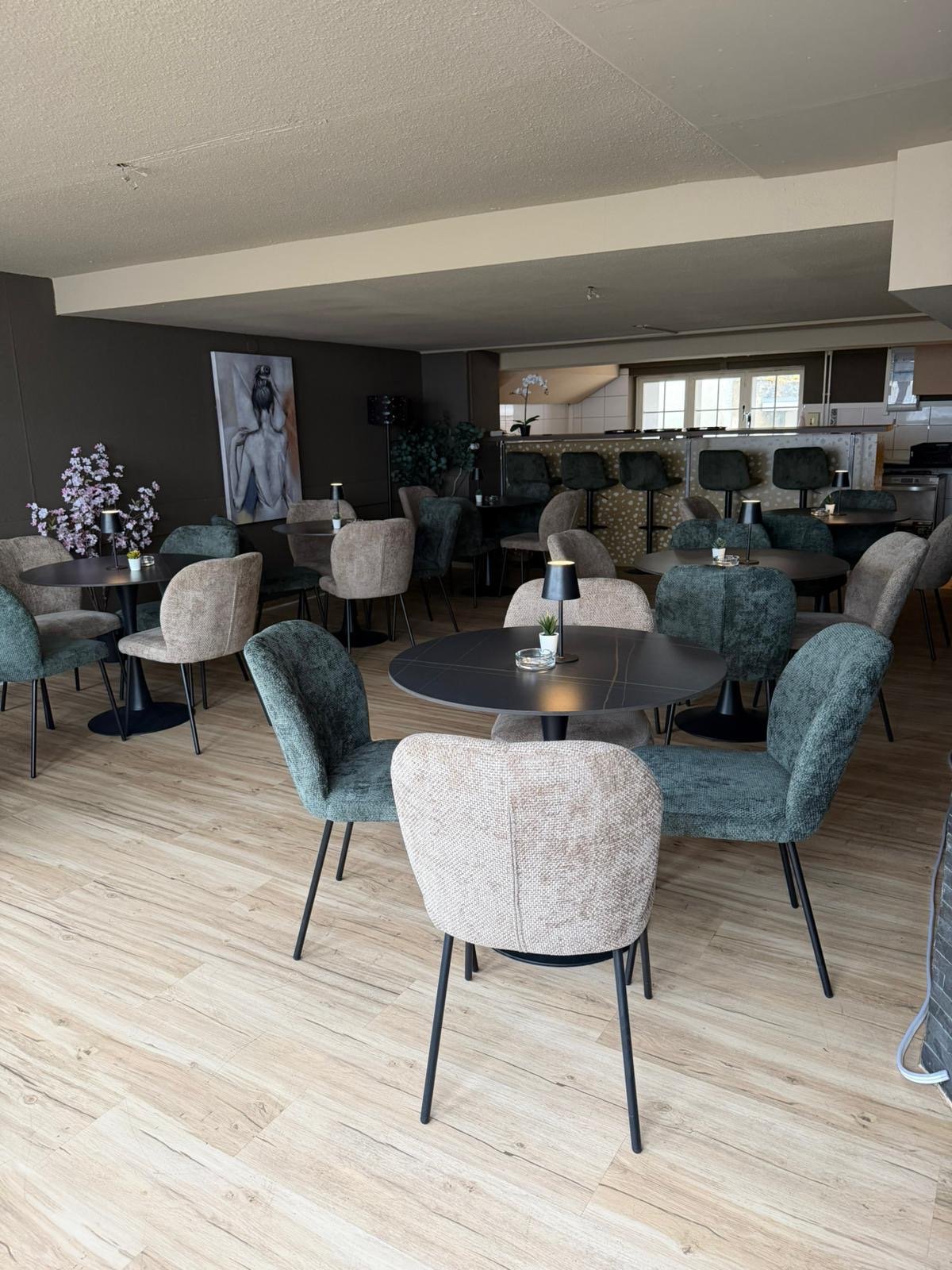 Modern indoor dining area with round tables and chairs, wooden floor, round lamps on tables, decorative plants, bar counter with chairs, and artwork on the wall.
