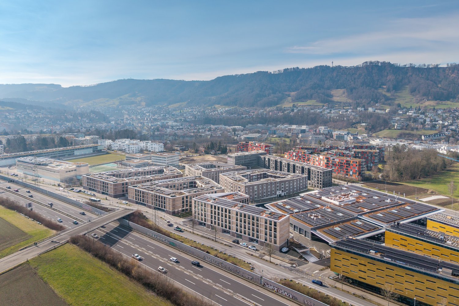 Aerial view of a city with a mix of residential and commercial buildings, highways, and parking lots. The city is surrounded by hills and mountains in the background, creating a scenic landscape.