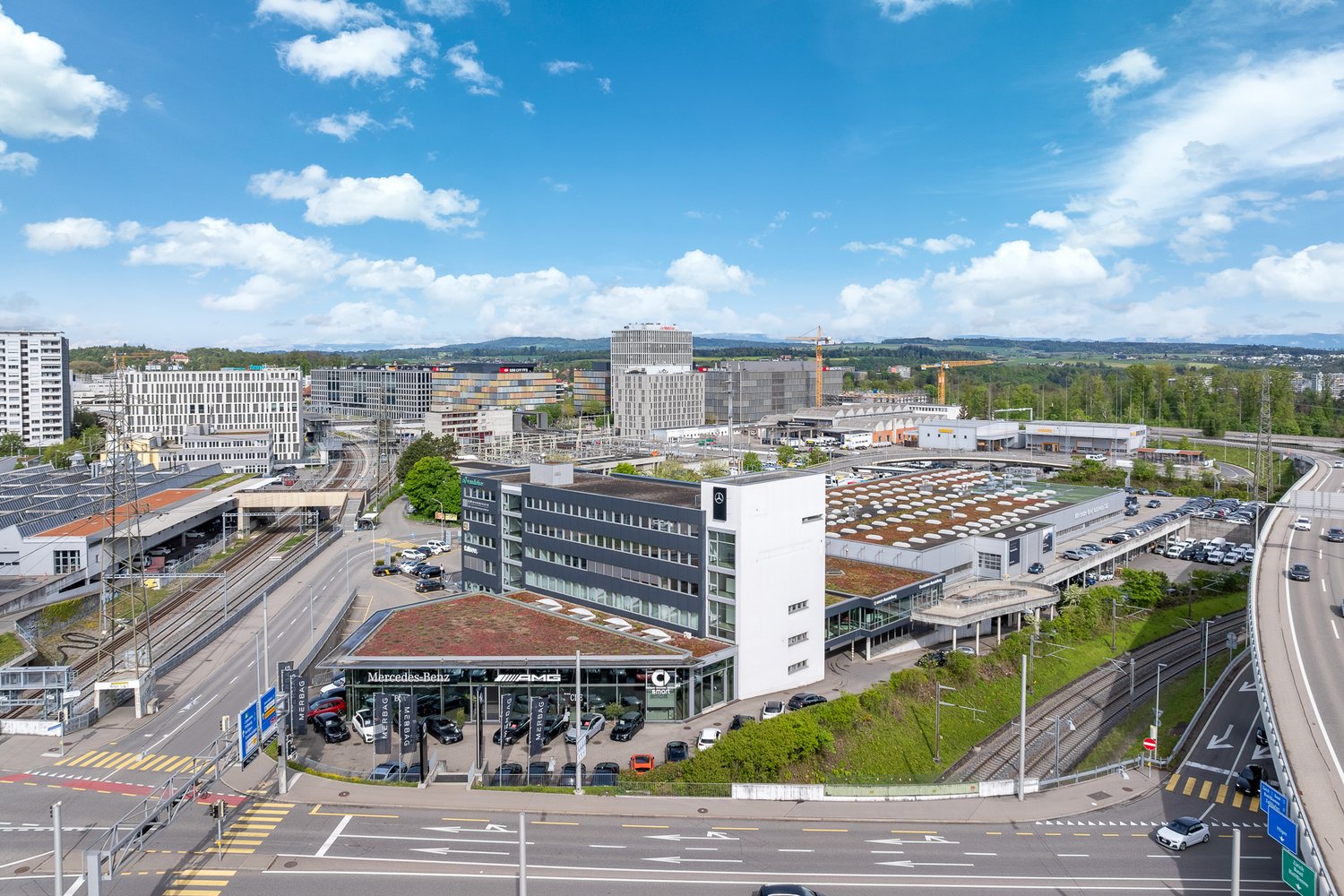 modern commercial buildings, parking lot with multiple cars, nearby train tracks, urban landscape with trees and mountains in the distance, multi-level structures, well-maintained green spaces, visible road network with cars