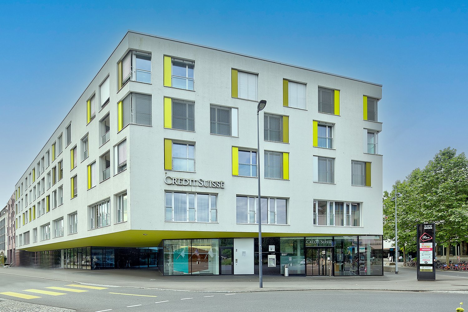 A modern, multi-story commercial building with a gray exterior and yellow accents. The building has large windows and a prominent Credit Suisse logo displayed on the facade. There are retail spaces on the ground floor and offices or apartments on the uppe