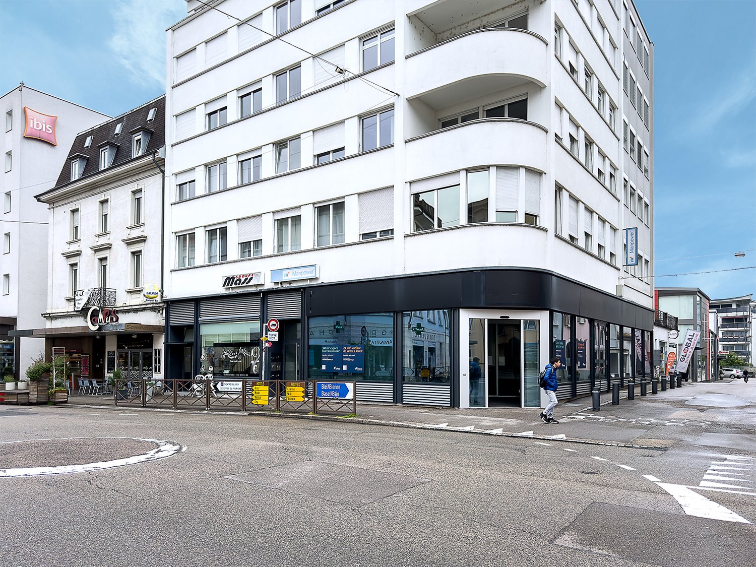 High-rise building, glass windows, black and white facade, store fronts, cafe with chairs, man walking on the sidewalk, greenery in front