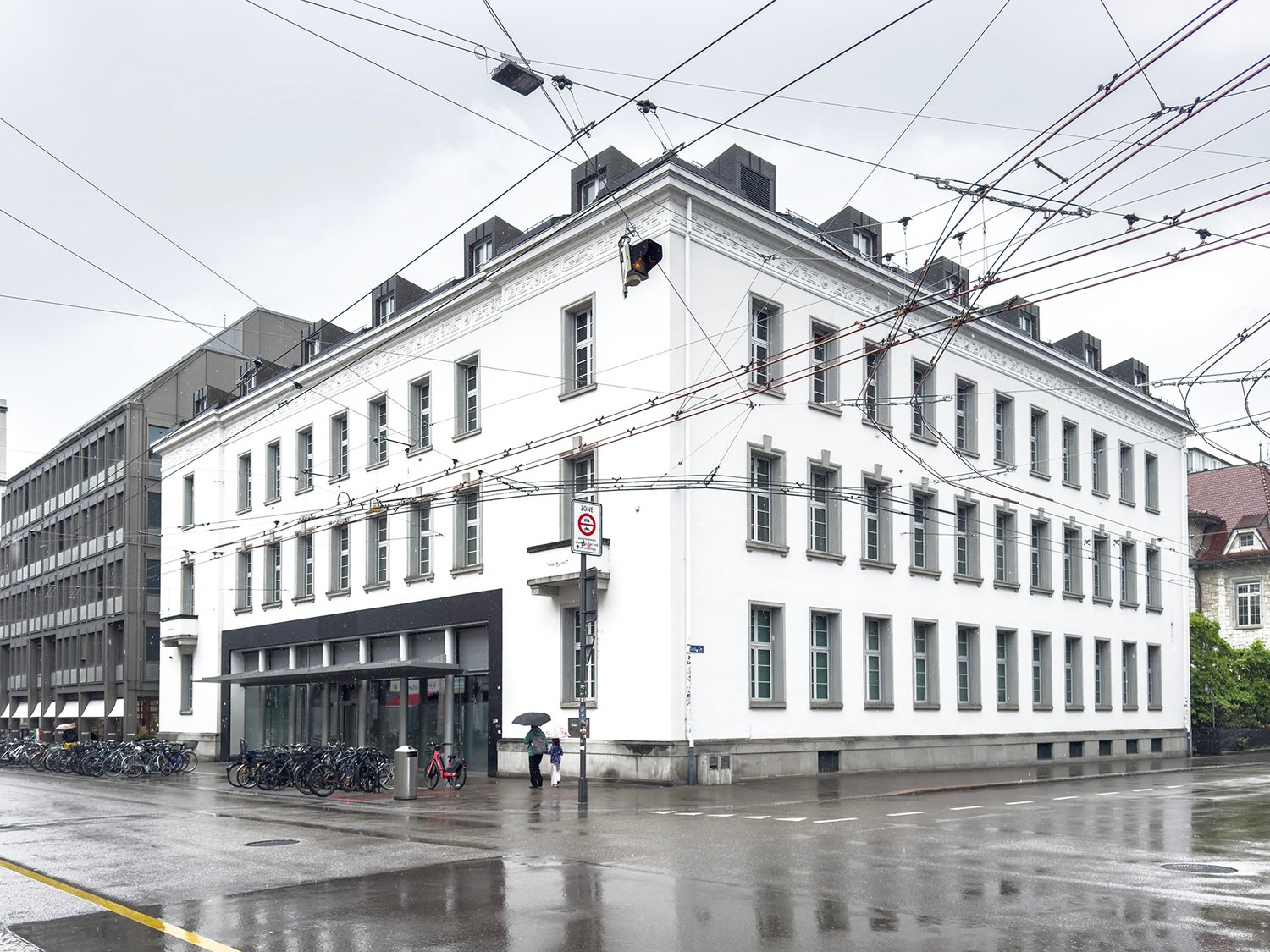 A multi-story white building with ornate architectural details, including windows and a prominent roofline. The building appears to be located in an urban area, with power lines and other infrastructure visible overhead. There are bicycles parked outside