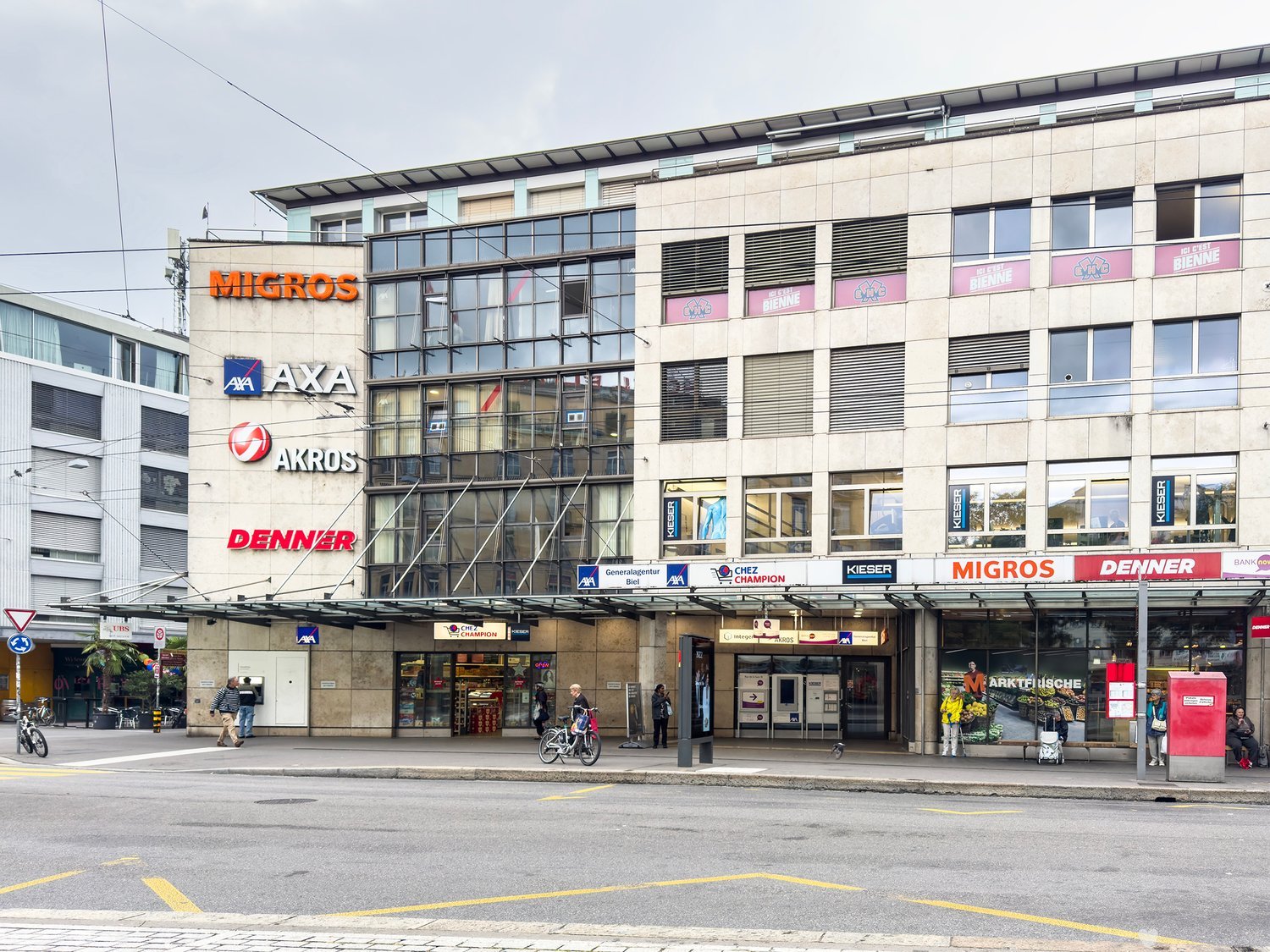 Multi-story commercial building with various retail stores and businesses, including Migros, Axa, Akros, and Denner. The building has a modern glass and concrete facade with signage for the different companies. There are parking spaces in front of the bui