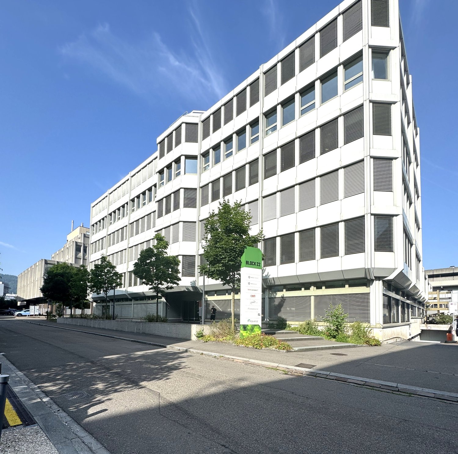 Multi-story office building with a modern, angular design featuring large windows and louvered facades. The building has a prominent green sign labeled 'Block 22' on the ground floor. There are trees and landscaping around the building, and a paved road i
