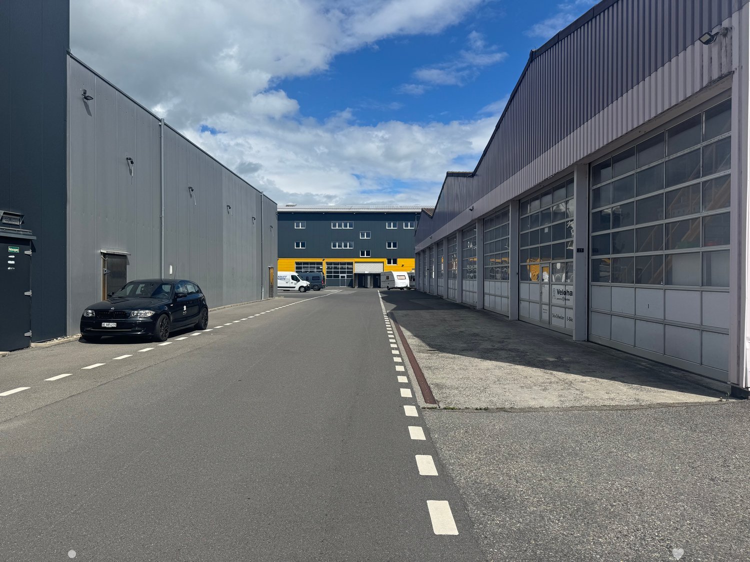Industrial complex with two large buildings, an empty road between them, multiple windows, a black car parked on the left side, and several vehicles parked near the entrance.