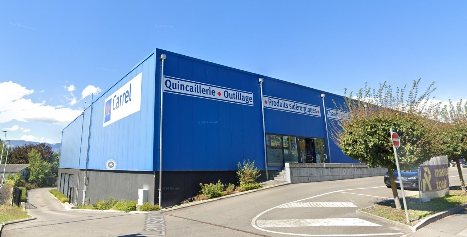 Large blue and white building with the sign 'Carrel Quincaillerie Outillage' indicating it is a hardware and tools store. There is a parking area in front of the building.