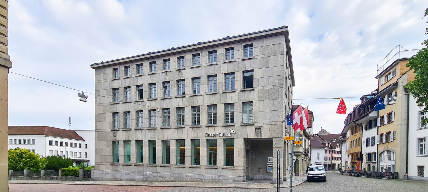 A commercial building with glass windows, a brick front, and a large sign reading 'Credit Suisse'. A white van is parked in front.