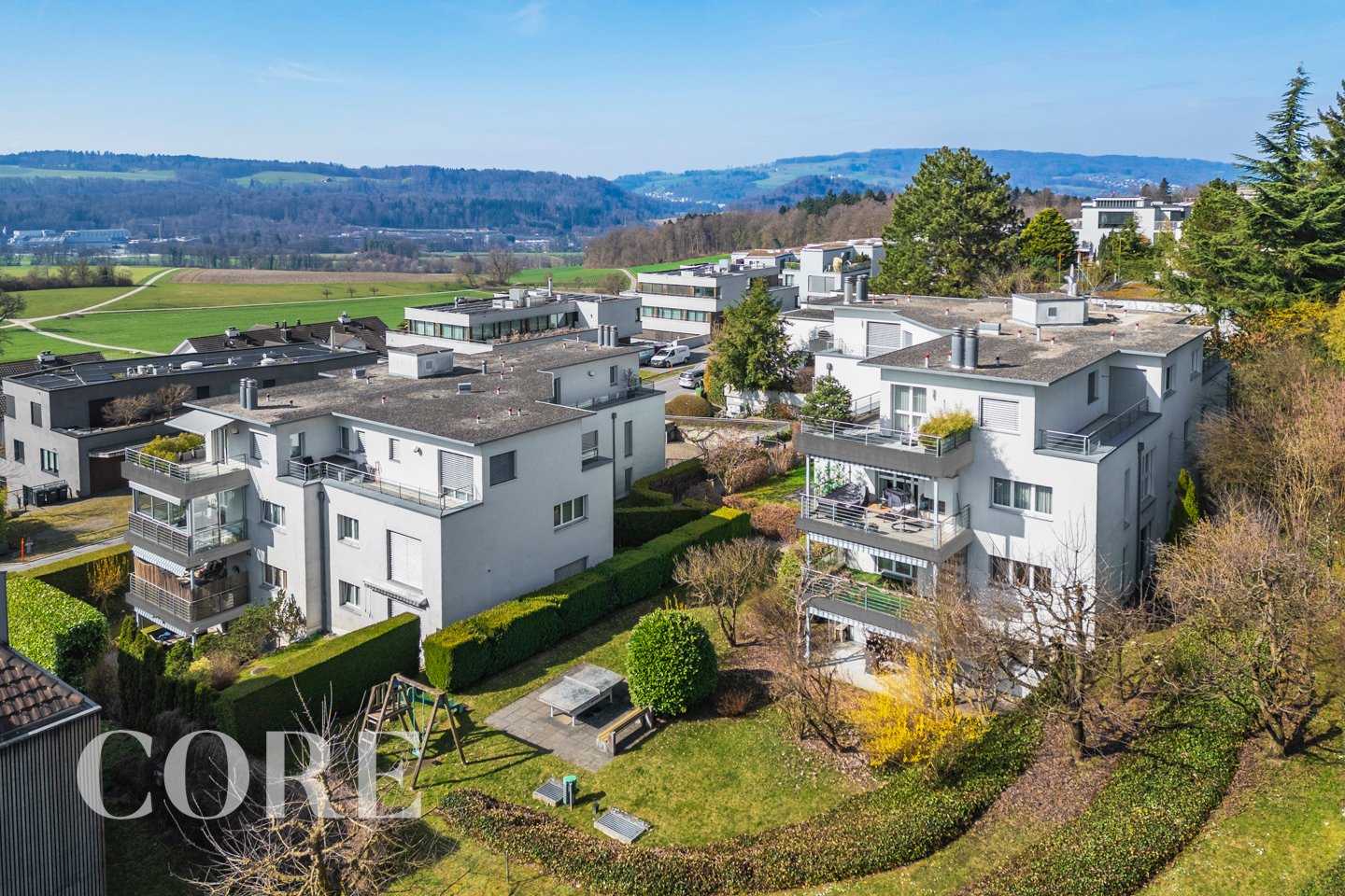 Aerial view of a residential area with multiple white buildings, each with multiple floors and balconies, surrounded by greenery and trees, with a clear view of the surrounding landscape.