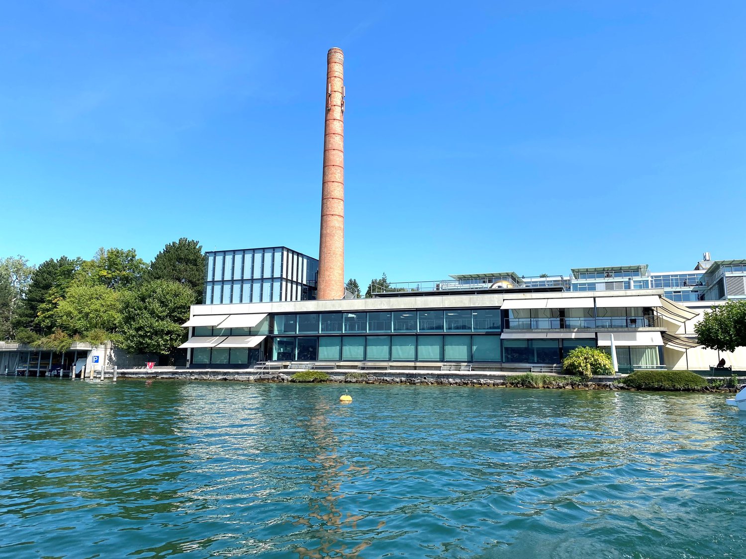 modern architectural building with glass windows and a tall chimney, set on a waterfront with a boat visible