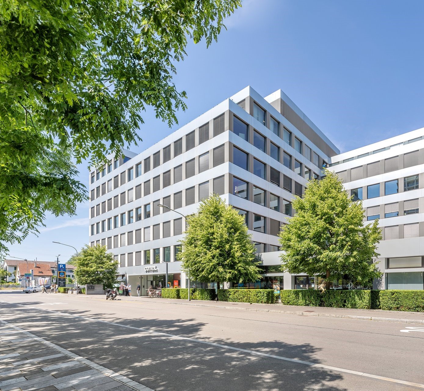 Raised ground floor, office building, several floors, many windows, hedges and trees in front, street with parked car