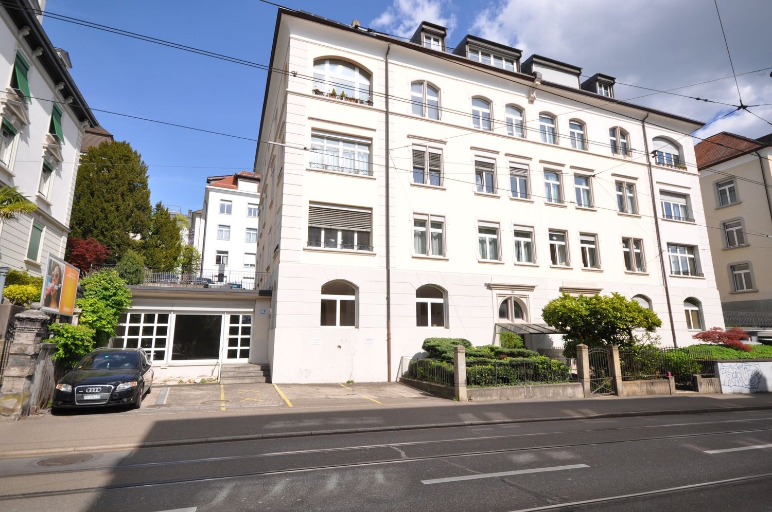 4 story building, white exterior, multiple windows, car parked in front, street with tram tracks