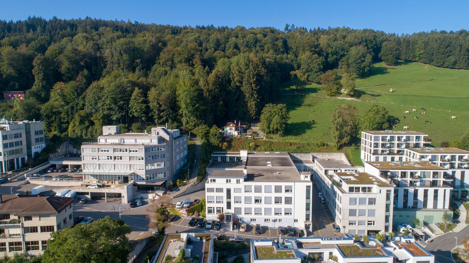 The image shows an aerial view of a town surrounded by a forested, hilly landscape. The town consists of several multi-story office and residential buildings, with a parking lot and some smaller buildings in the foreground. The buildings have a modern, co
