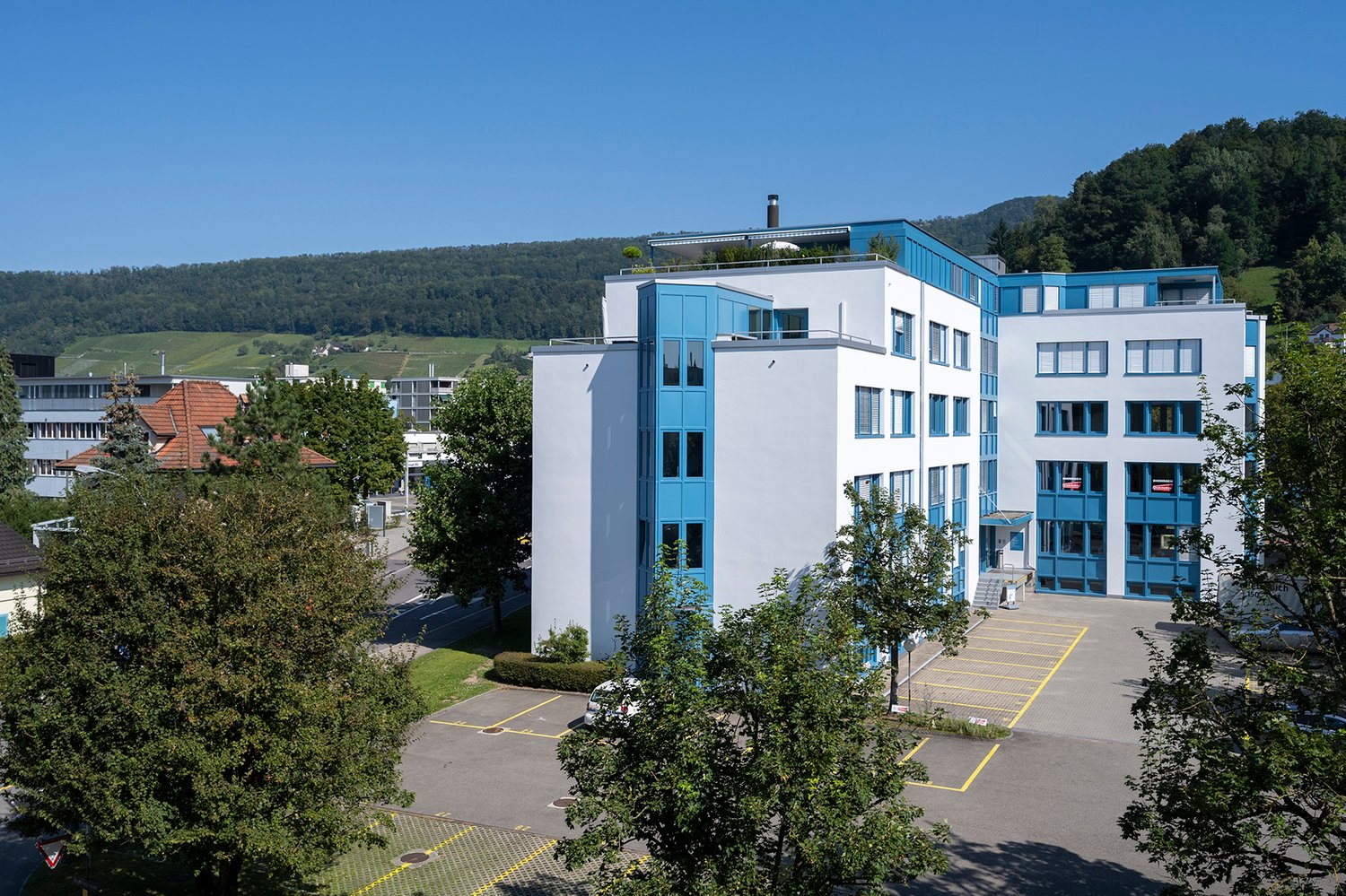 modern multi-story building, white exterior, blue accents, parking lot, scenic mountain view, several windows, entrance with steps