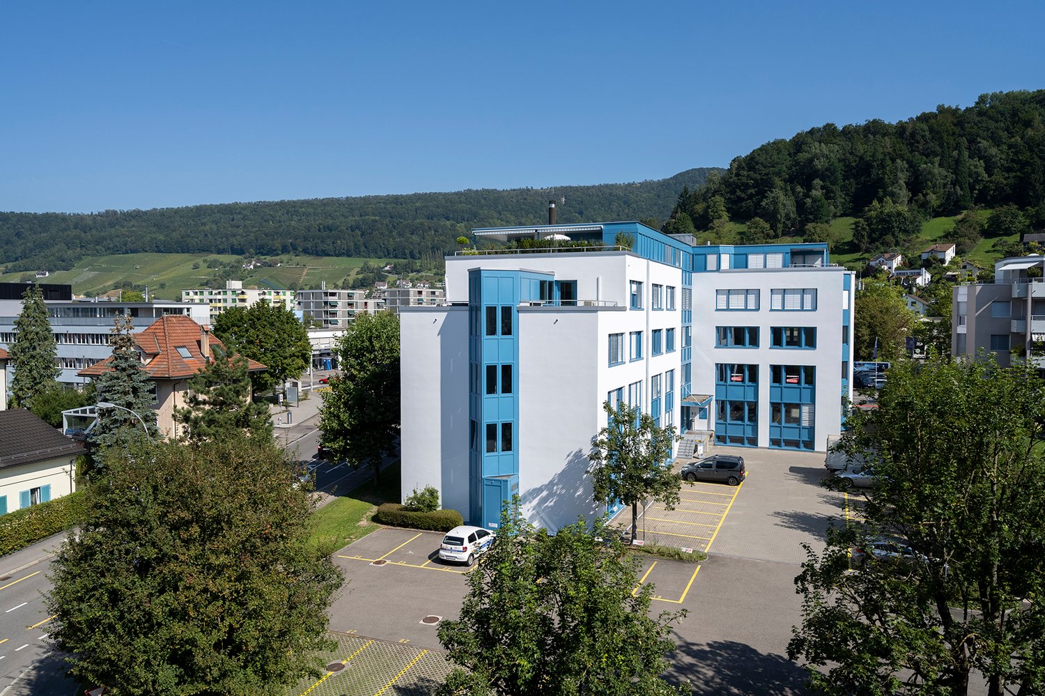 Multi-story office building with white and blue exterior, surrounded by trees and greenery, with a parking lot in the foreground