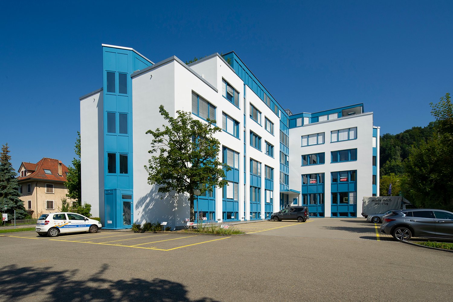 White and blue multi-story building with multiple windows, entrance doors, and parking spaces.