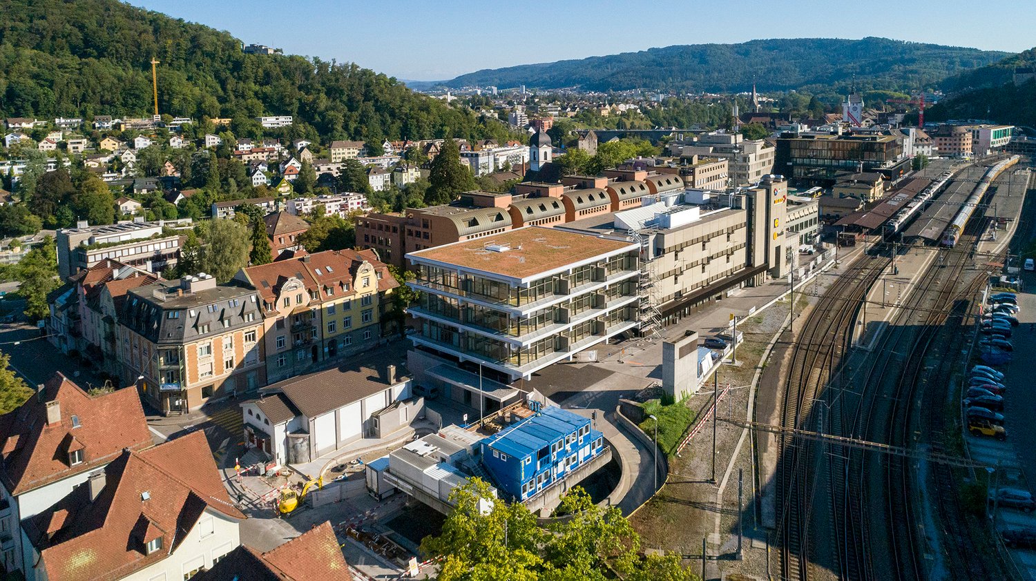 Aerial view of a cityscape with various buildings, trees, and a railway station in the foreground. The buildings are a mix of architectural styles, with some having red roofs. The trees are lush and green, and the railway station has several tracks and a