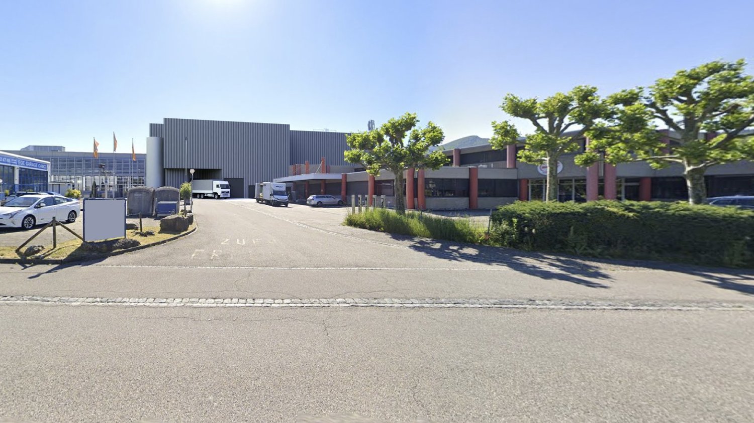 Industrial building with large parking area, multiple trucks and cars parked, trees and bushes in front