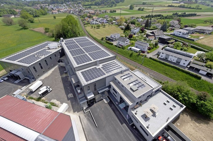 Large building with solar panels on the roof, multiple parking spaces, surrounded by houses and greenery