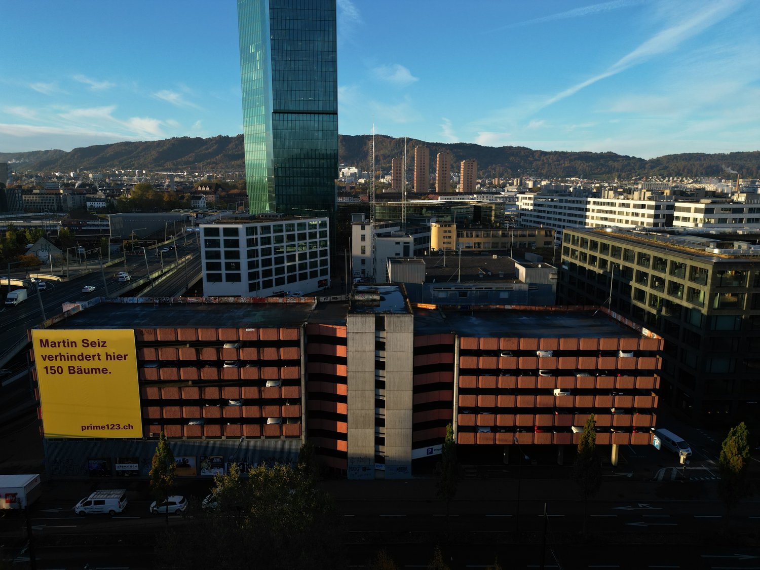 Tall glass office building, surrounded by smaller multi-story brick buildings, with a city skyline in the background