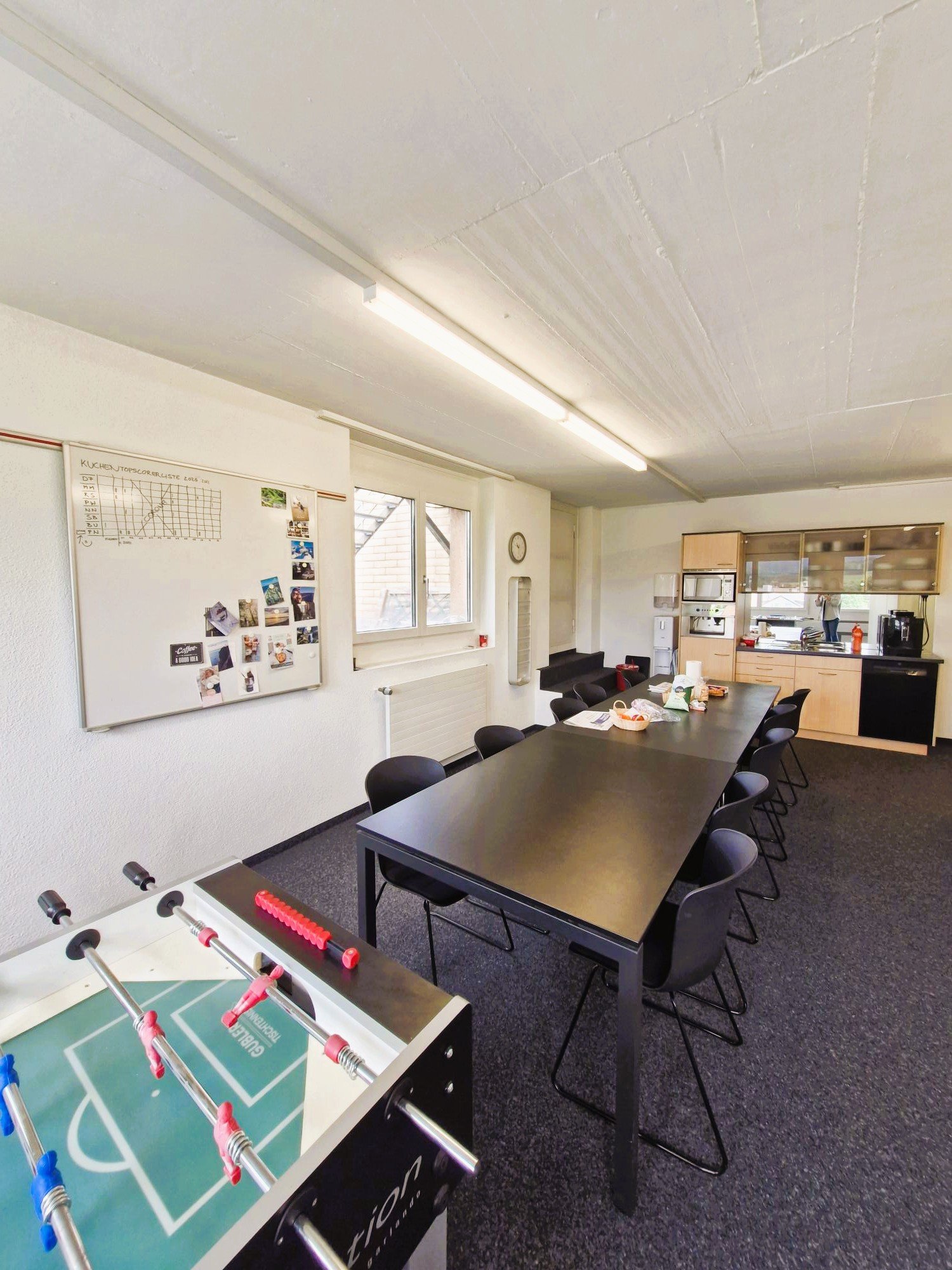 large room, long black table, chairs, whiteboard, table football, cabinets, shelves, clock, radiator, microwave, dishwasher, coffee machine