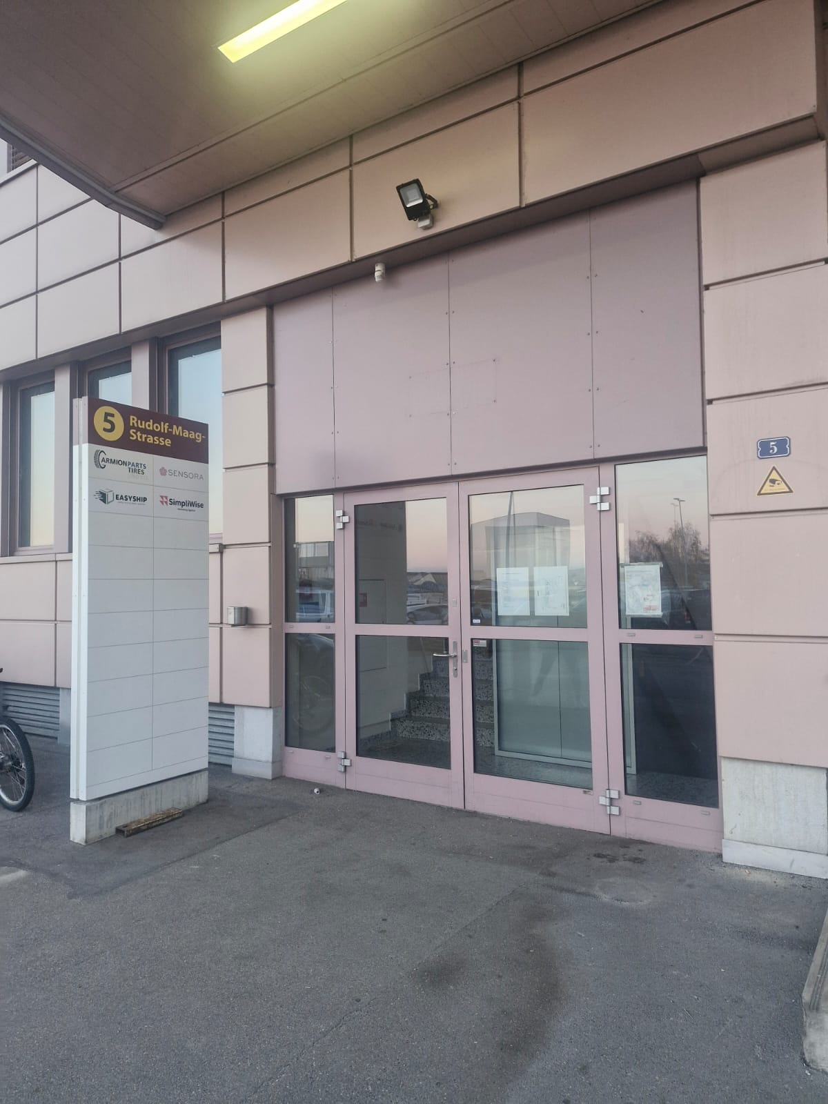 Large pink building entrance with triple doors, glass windows, security camera, illuminated ceiling light, white signpost, bicycle parking.
