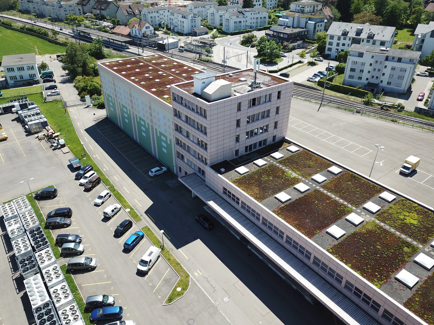 Aerial view of a building with rooftop green space and surrounding parking lot, numerous cars parked, greenery, and trees