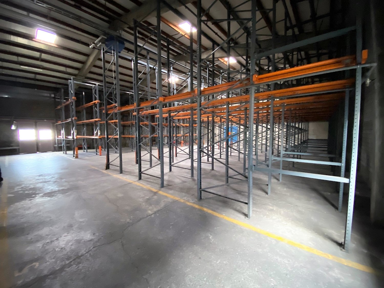 Empty warehouse with metal shelving units, concrete floor, yellow line on the floor, windows on the left side, metal beams on the ceiling