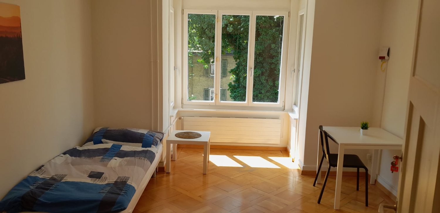 Spacious bedroom with large windows overlooking a garden, hardwood floors, a bed with patterned bedding, a small table, and a chair.