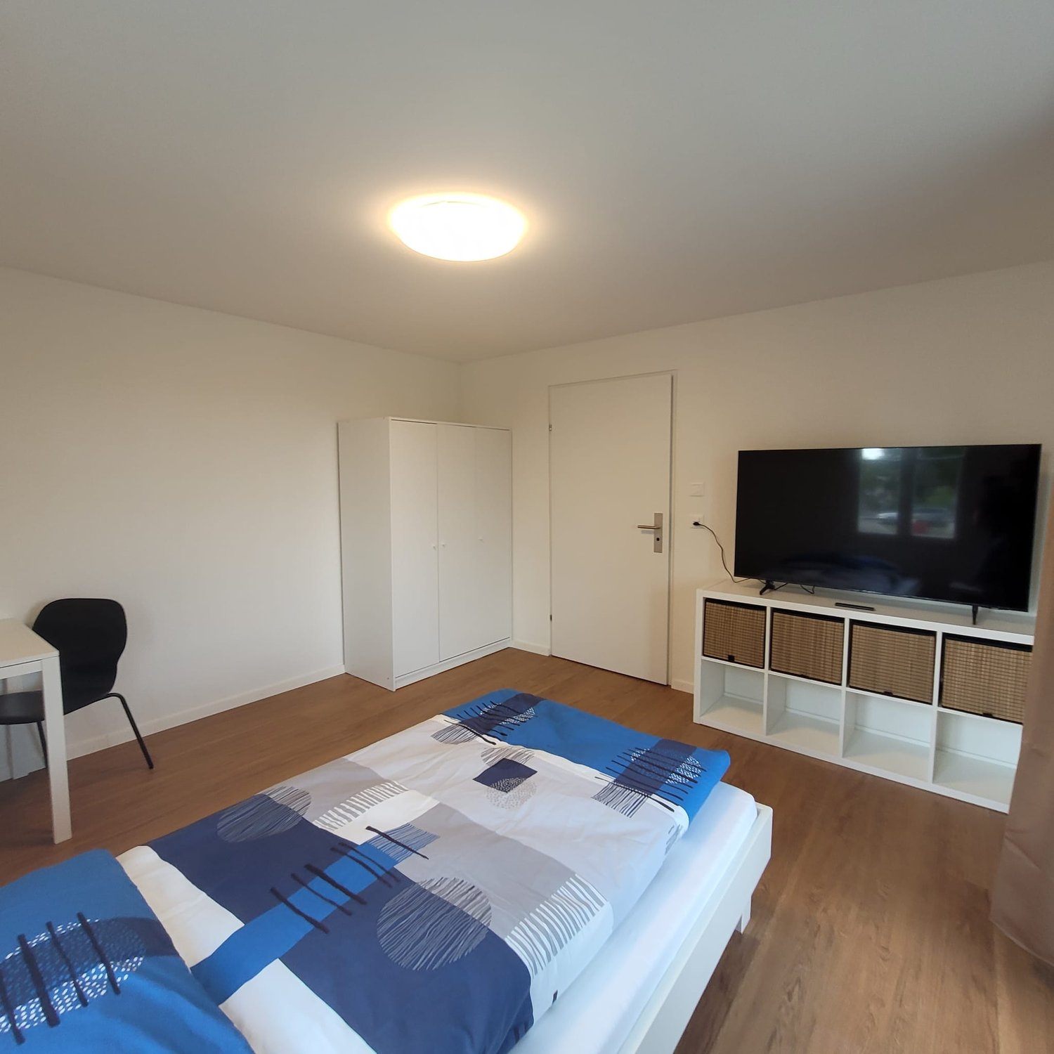 room with bed, wardrobe, tv, desk, chair, wooden floor, white walls, ceiling light
