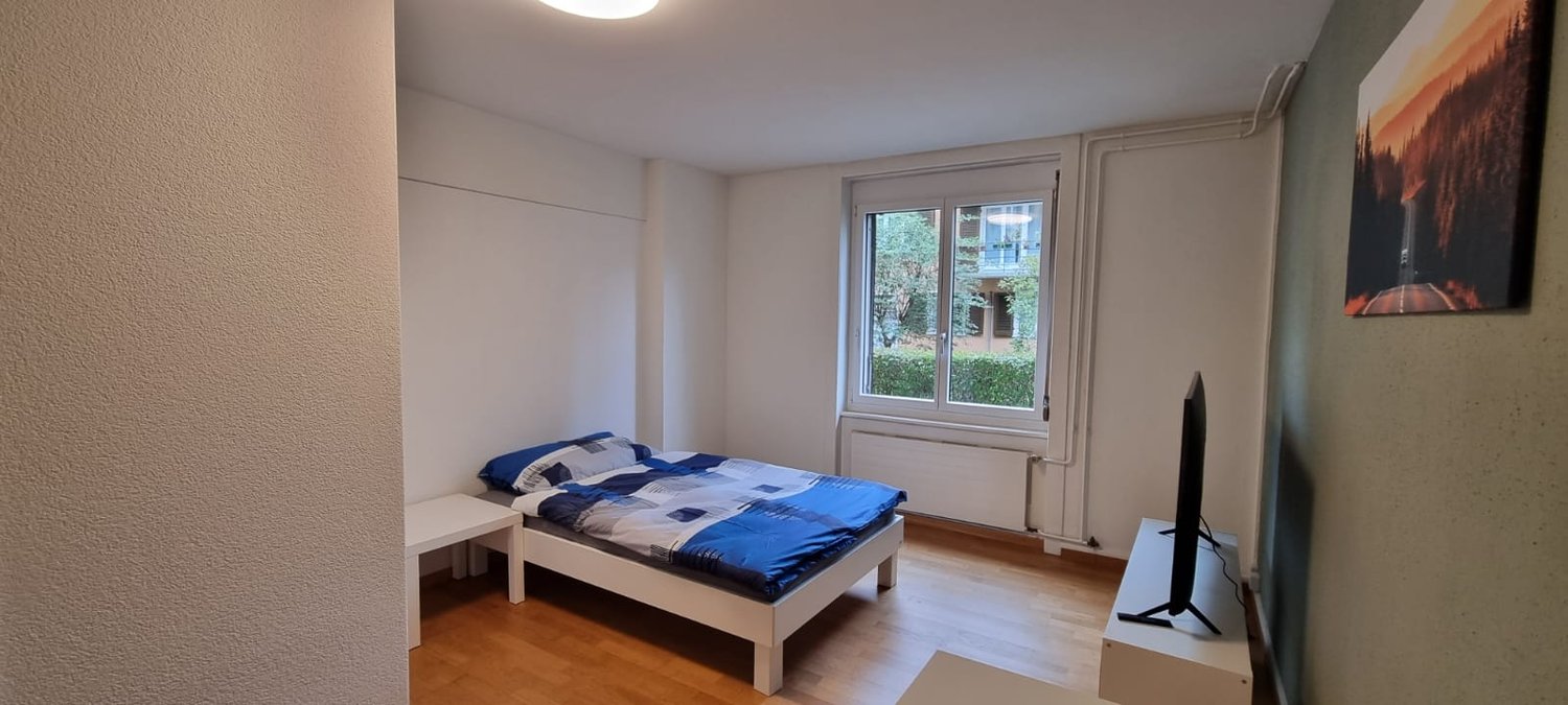 Single bed with blue and white bedding, nightstand with TV on top, white walls, wooden floor, large window with views of trees and a building outside