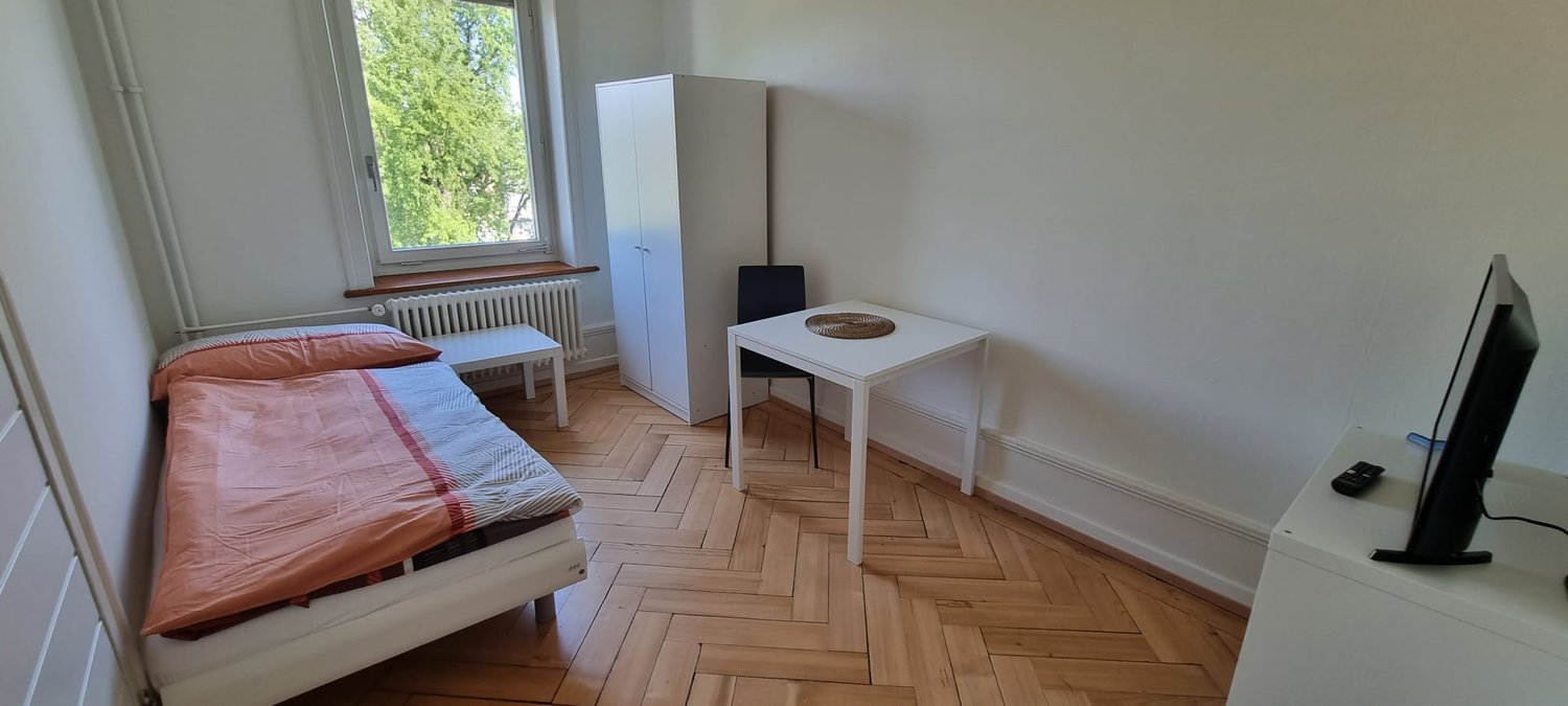 Bedroom with hardwood flooring, white walls, large window, bed, dresser, and small table