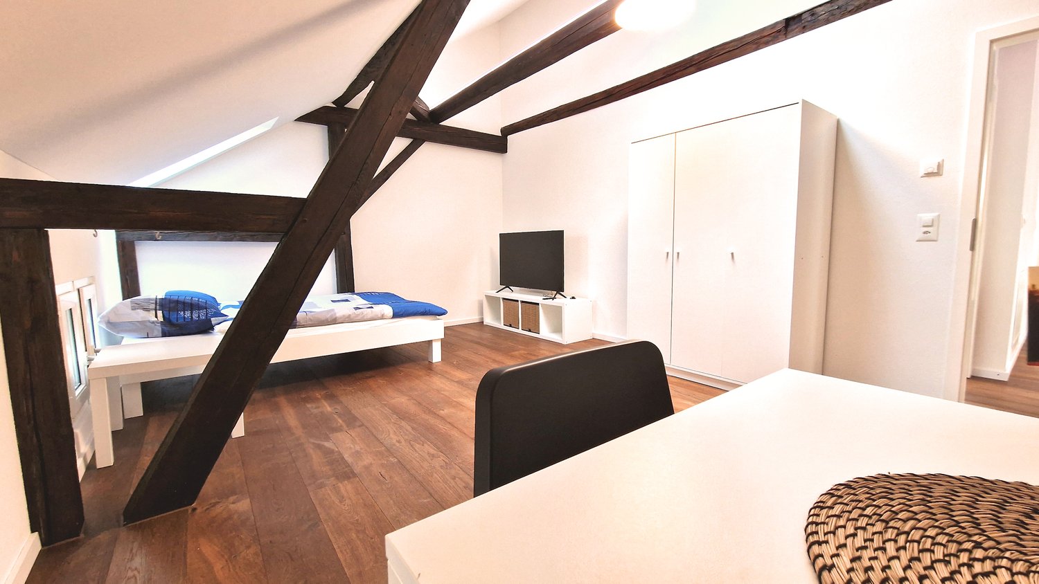 Bedroom with double bed, white wooden bedframe, dark brown wooden beams, white walls, TV, wooden floor, desk, and chair