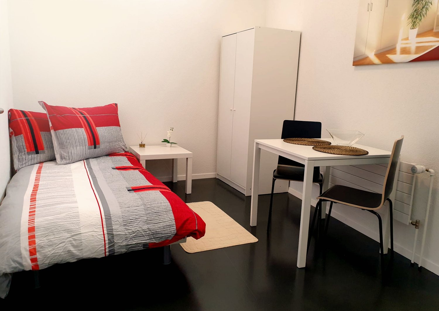 a small room with bed, desk, two chairs, white wardrobe, heater on wall