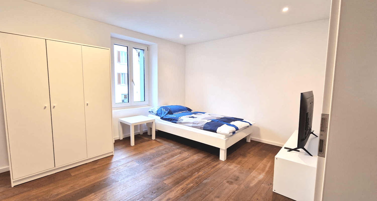 A single bed, a white night stand, a large white wardrobe, a flatscreen TV and hardwood floors