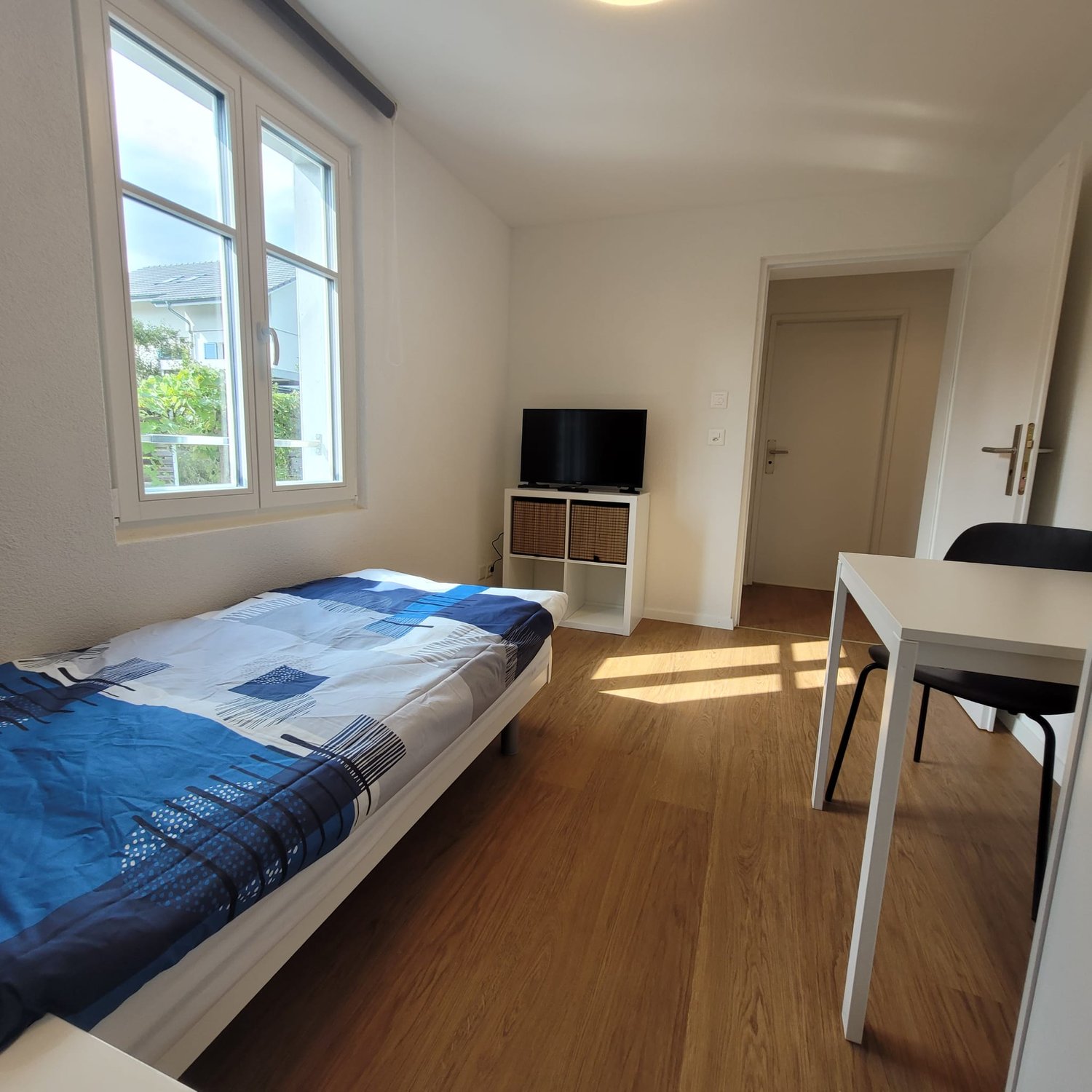 single bed, flat screen TV on shelf, desk, black chair, hardwood floor