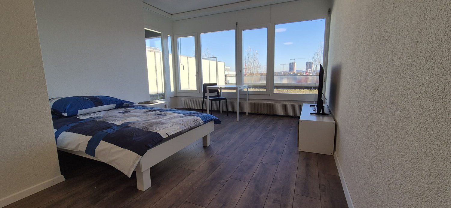Spacious bedroom with large windows offering a view, hardwood flooring, a white bed frame, and a TV stand.